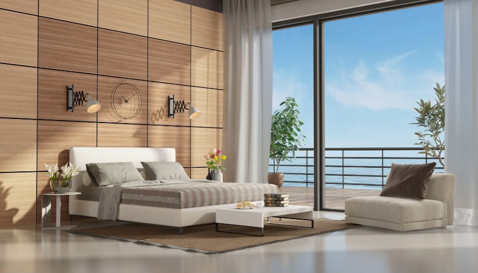 A modern bedroom with wood-paneled walls, a white bed, beige armchair, and a large sliding door revealing an ocean view.