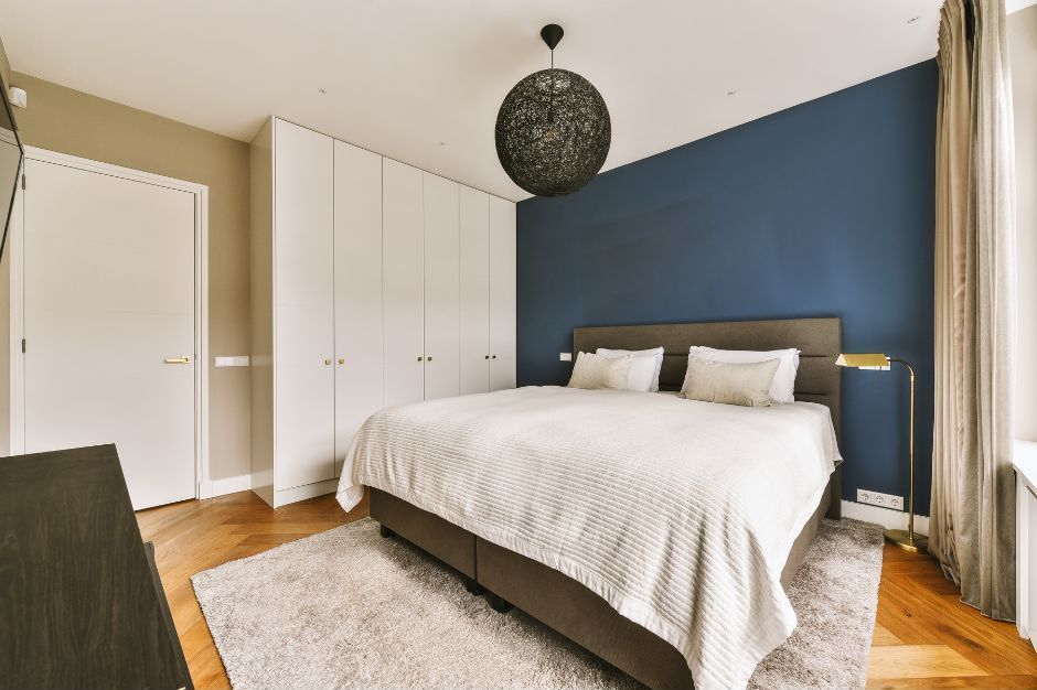 A bedroom featuring a large bed with a white comforter, set against a deep blue accent wall, with white wardrobes.
