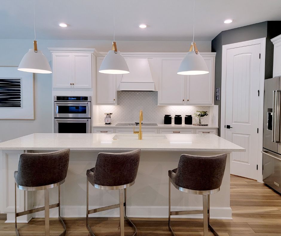 A modern white kitchen featuring an island with three gray bar stools, pendant lighting, and stainless steel appliances.