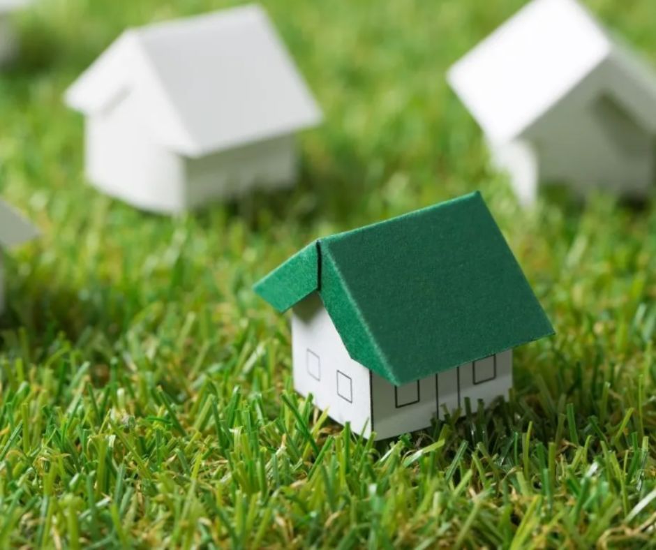 A small model house with a green roof sits in a patch of grass, surrounded by other white model houses in the background.