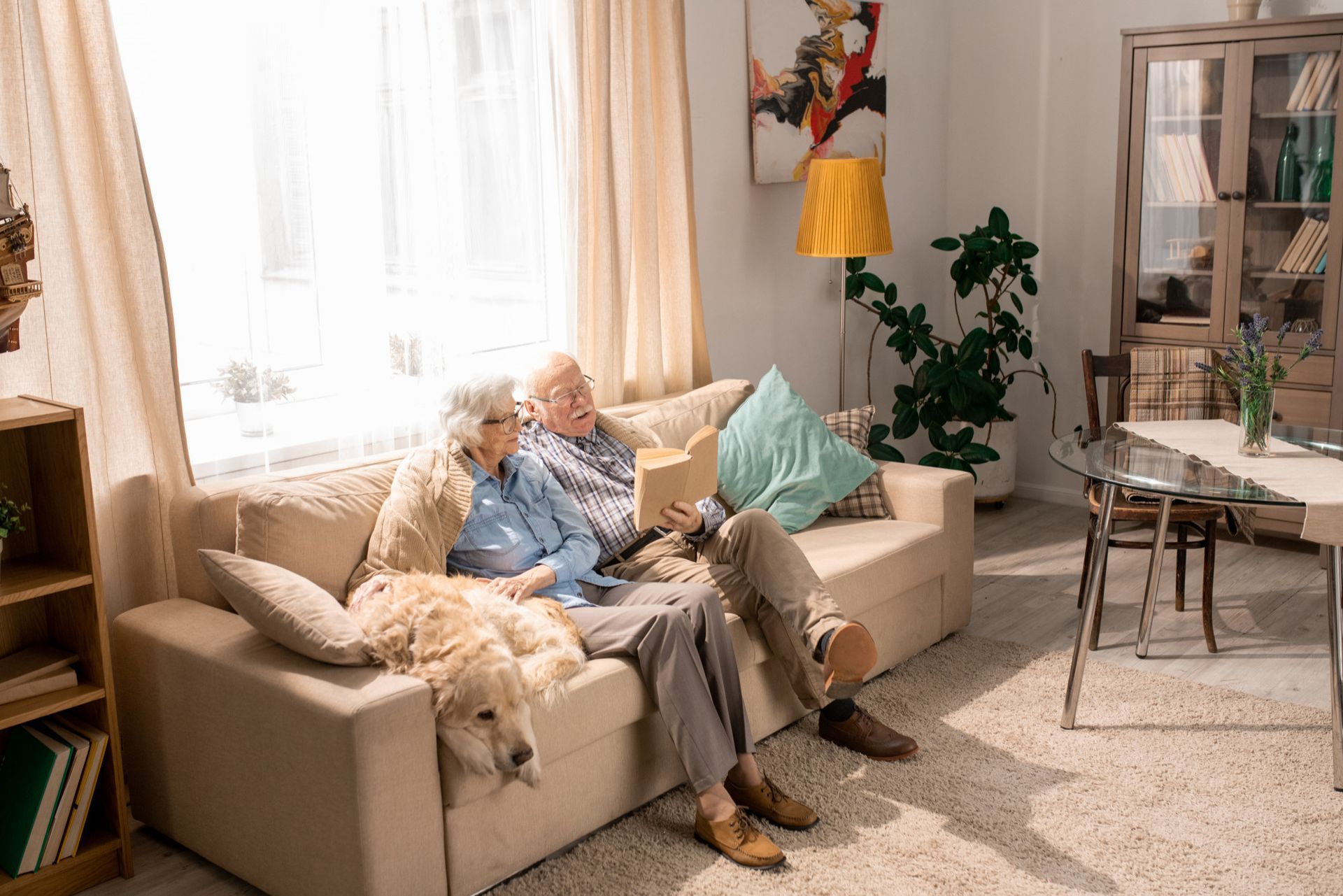 Two people sit on a beige couch with a dog, one reading a book, in a brightly lit living room with a green plant.