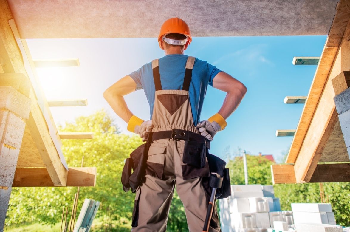 Construction worker standing with hands on hips beneath a house frame, wearing an orange hard hat and overalls.