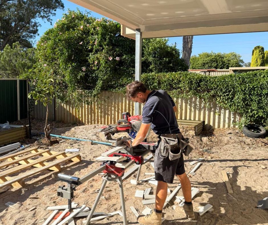 A person uses a miter saw on a stand in a sandy outdoor construction site, surrounded by wood scraps and yard fencing.