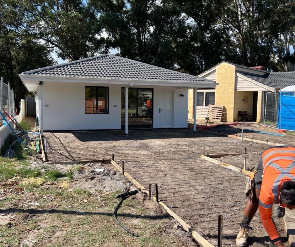 A worker in high-visibility gear prepares a dirt area for concrete construction in front of a modern residential home.