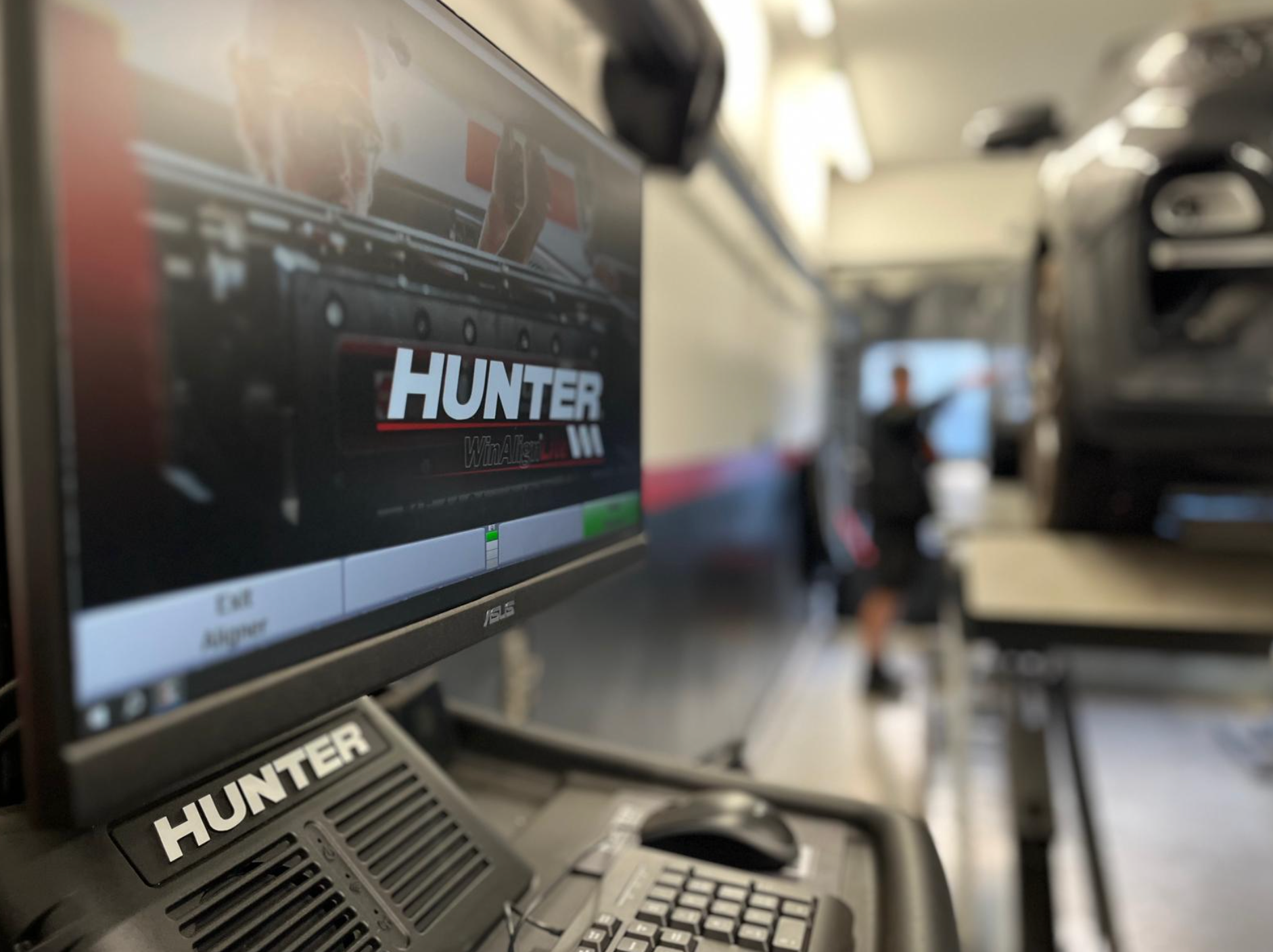 Hunter wheel alignment machine in a car repair shop. Car is on a lift, worker in background.