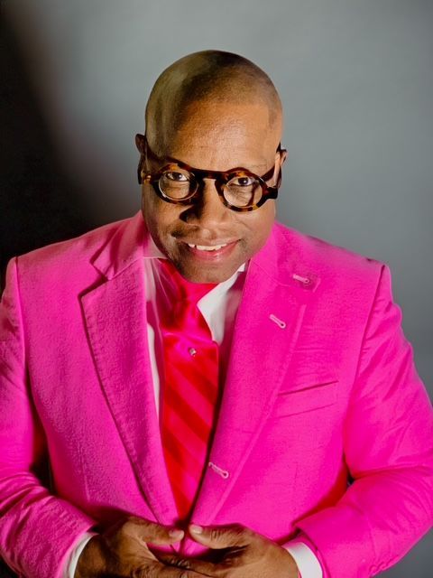 Man in a pink suit, red tie, and large glasses, smiling. Against a gray background.