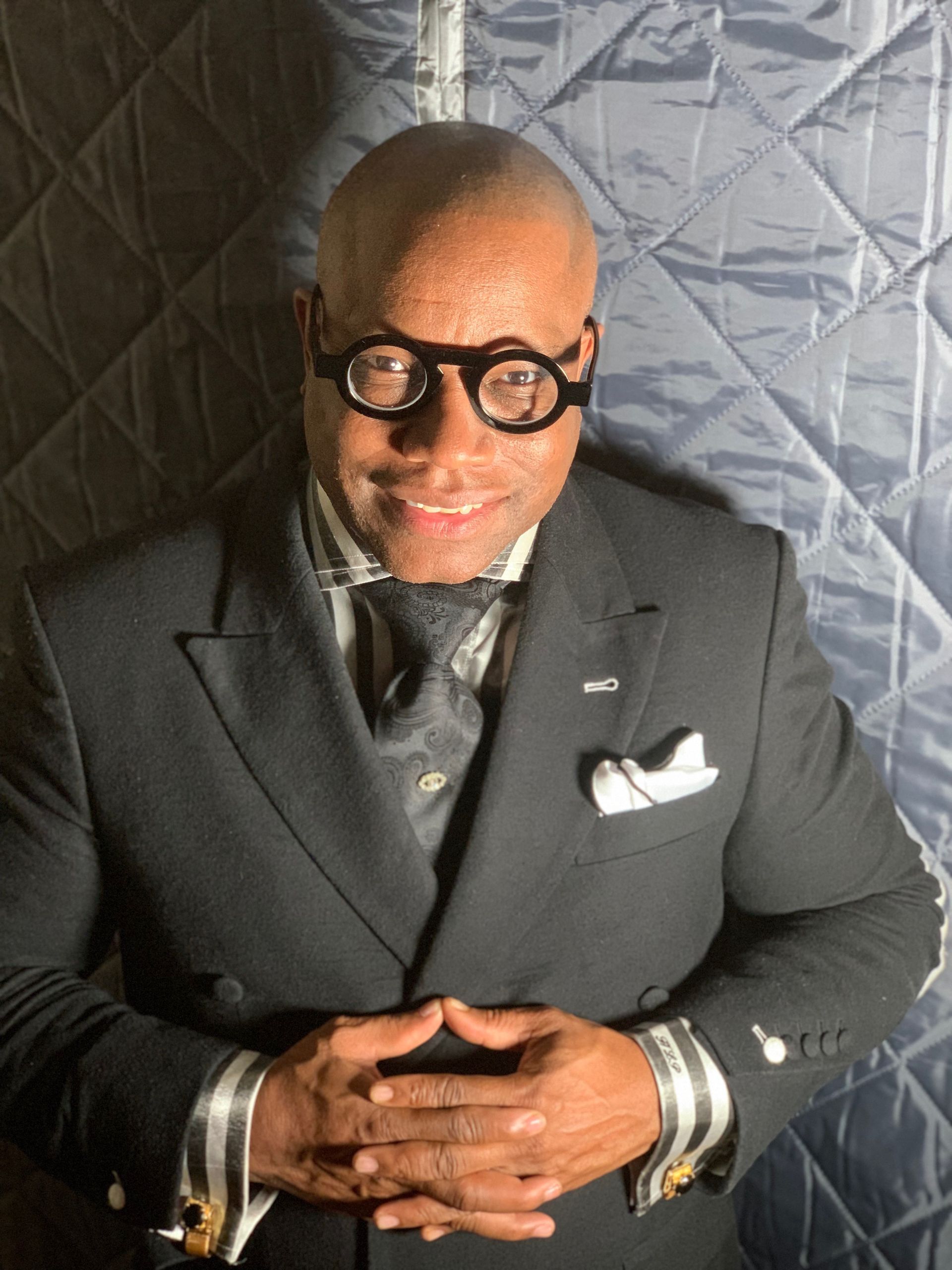 Man in a dark suit and round glasses, smiling. Hands clasped in front, against a textured backdrop.