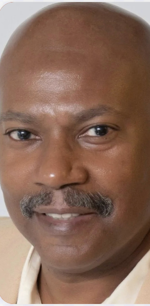 Close-up of a Black man with a bald head and mustache smiling. He wears a light-colored shirt.