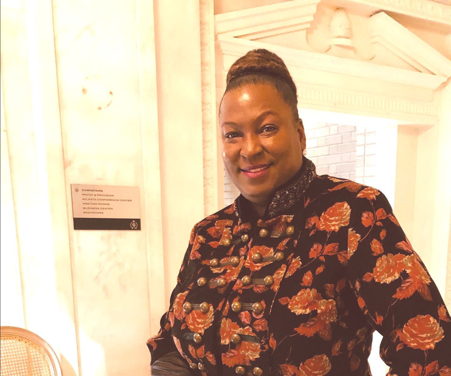 Woman in a black and orange floral jacket, smiling in a well-lit room with a wall sign.