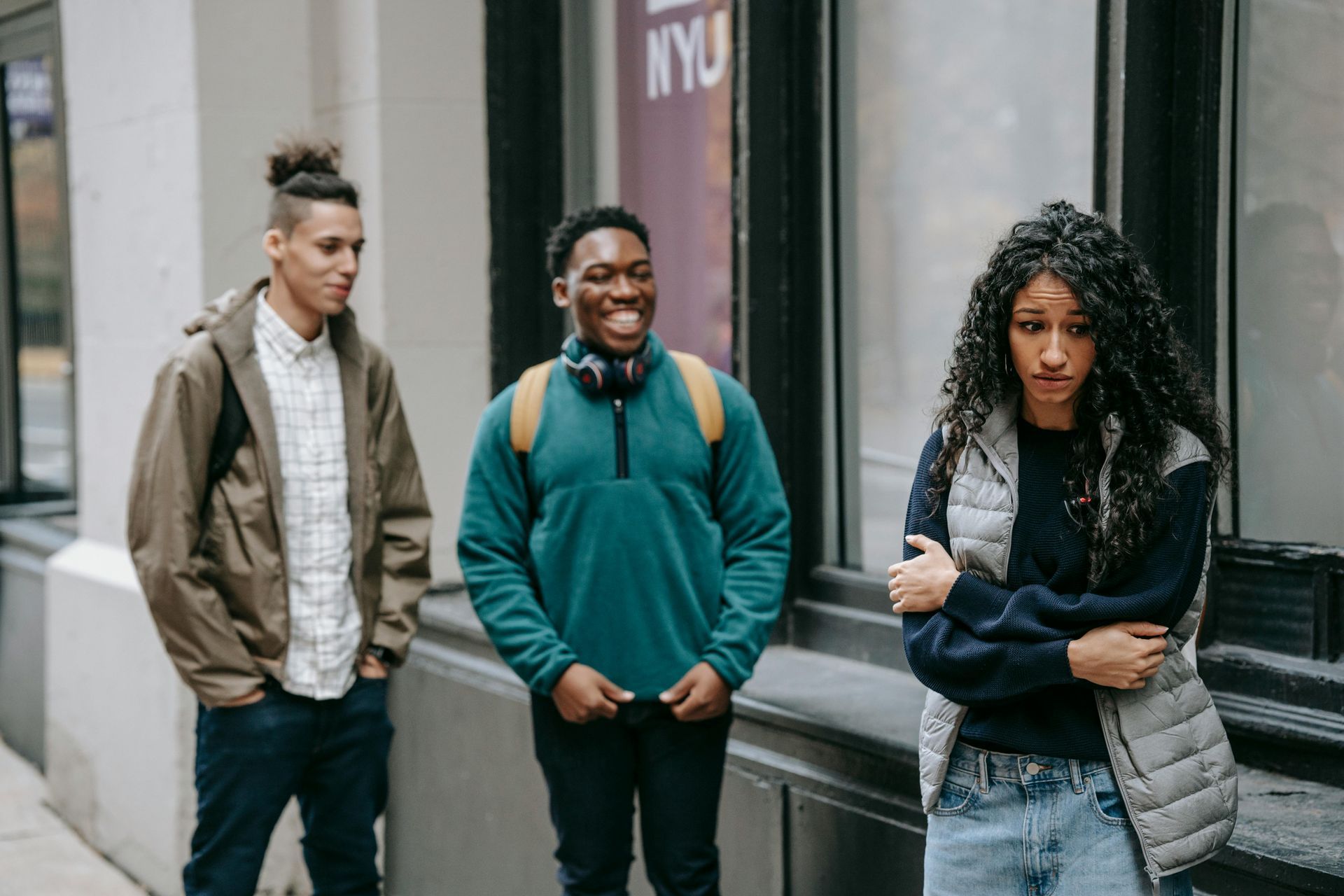 Three young people outside. Two men smiling, one woman looking unhappy, arms crossed.