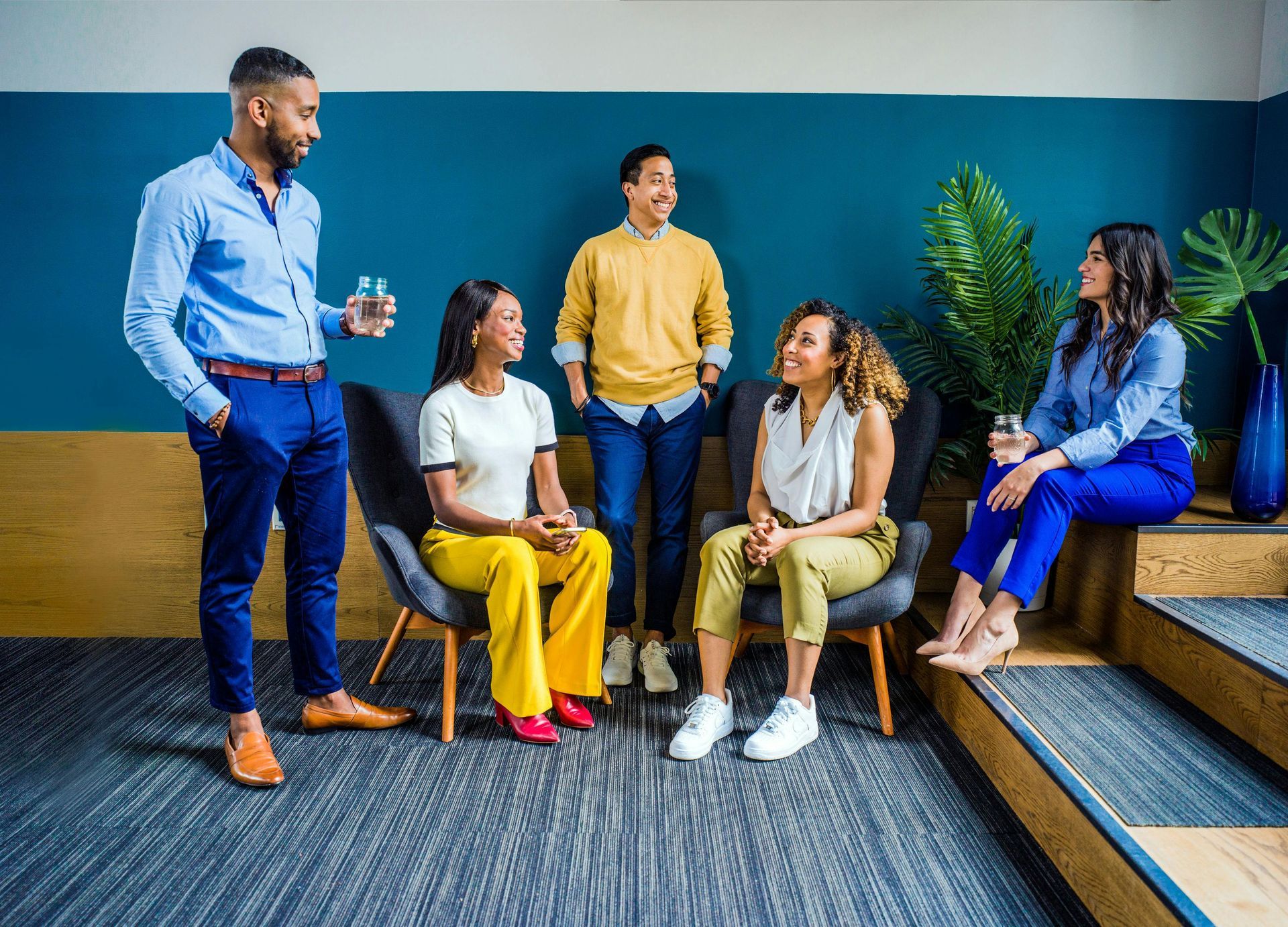 Five diverse people in a room, conversing. Some seated in chairs, others standing, smiling, in various colorful outfits.