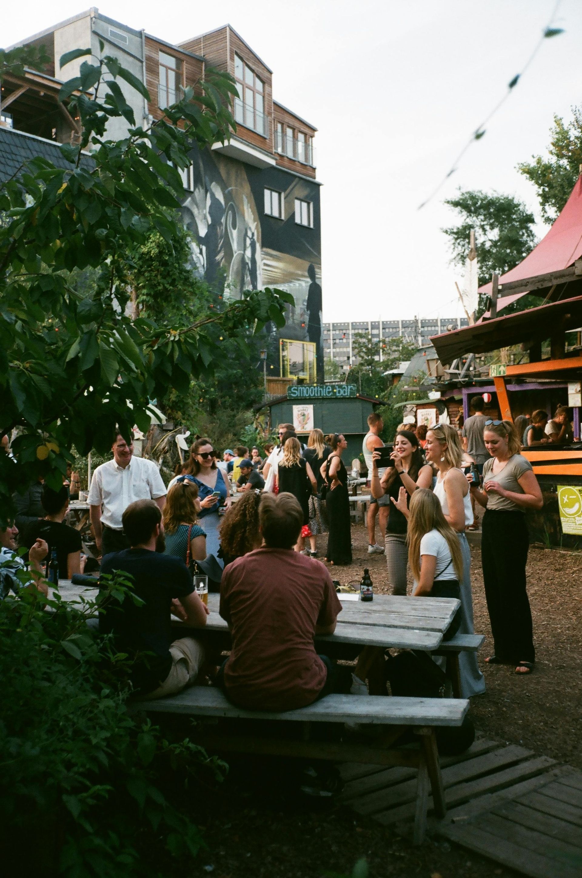People gathered outdoors, near picnic tables and food stalls. Evening, with a large mural on a building in the background.