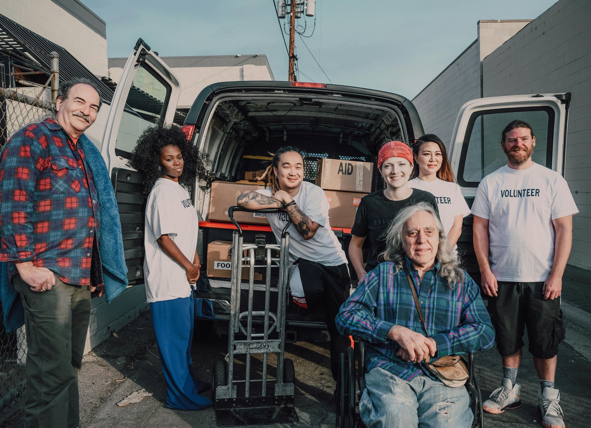 Group of people, including a person in a wheelchair, near an open van filled with boxes and a hand truck.