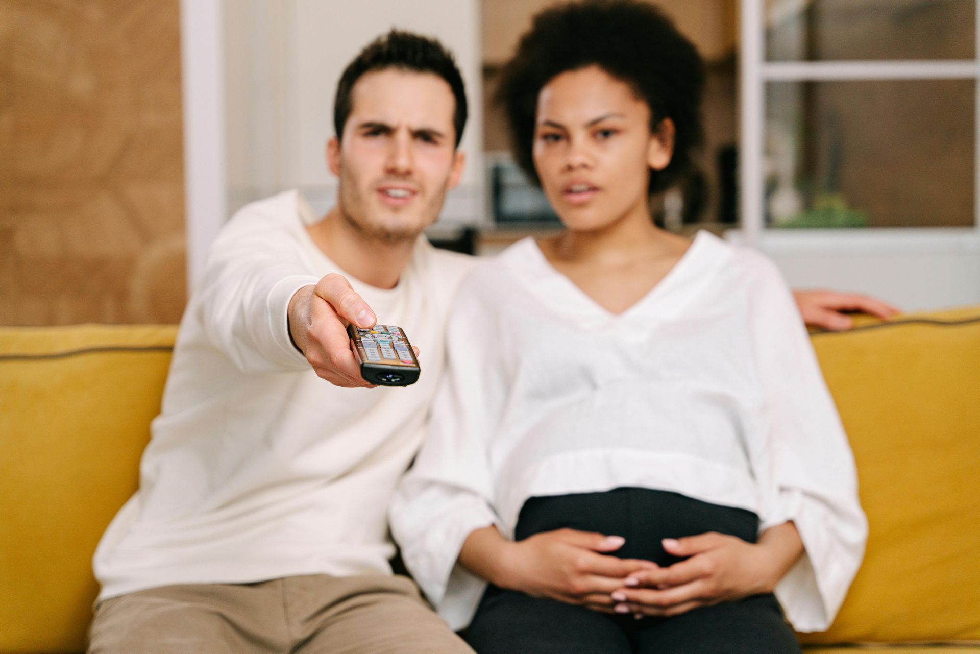Man holding remote, pregnant woman, sitting on yellow couch; focused, looking at TV.
