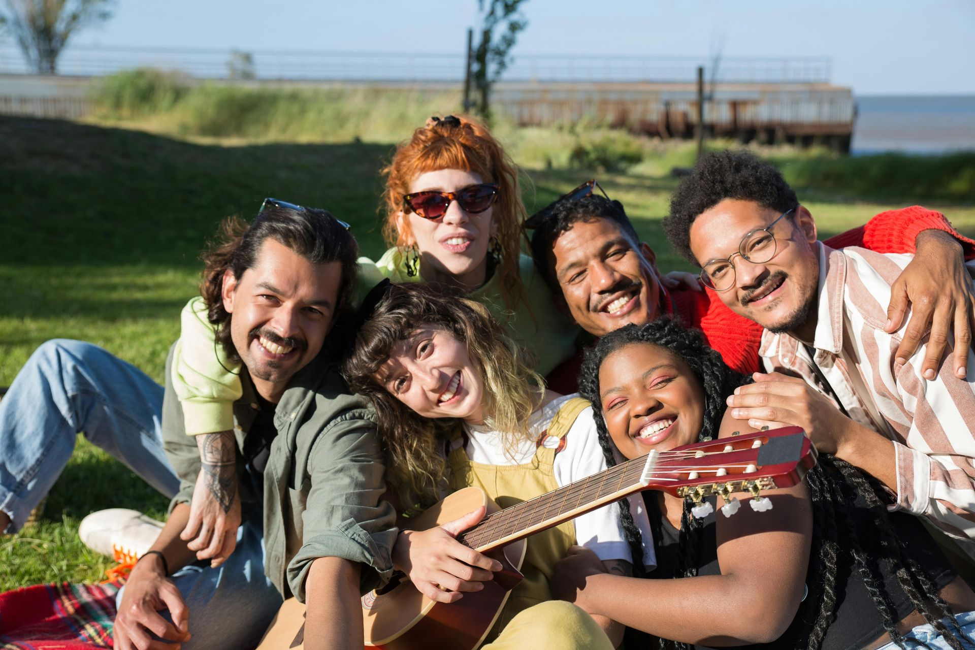 Group of six diverse friends smiling for a photo outdoors in a park, holding a guitar.