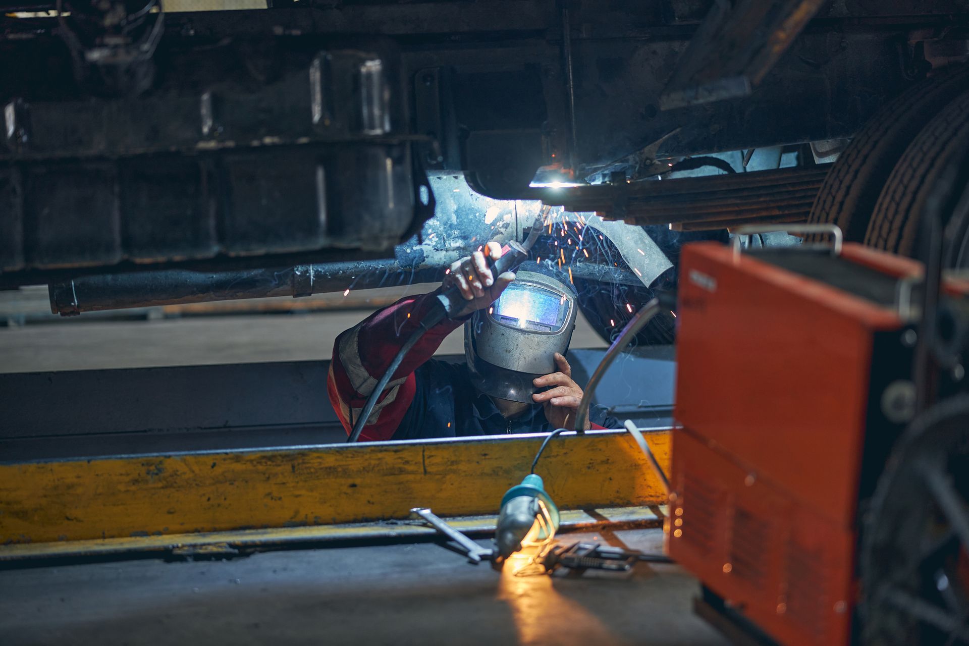 Welder in a workshop, welding under a vehicle with sparks flying.