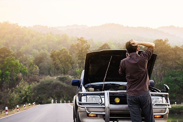 Person standing by a car with open hood on a mountain road at sunrise | Jeff's Automotive, Inc