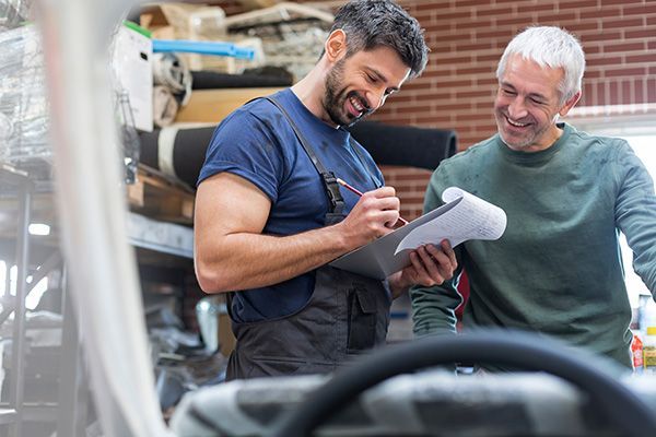 Two men reviewing a clipboard in a workshop, one smiling while writing notes. | Jeff's Automotive, Inc
