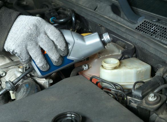 Gloved hand pouring fluid into a car’s engine bay from a silver bottle | Jeff's Automotive, Inc