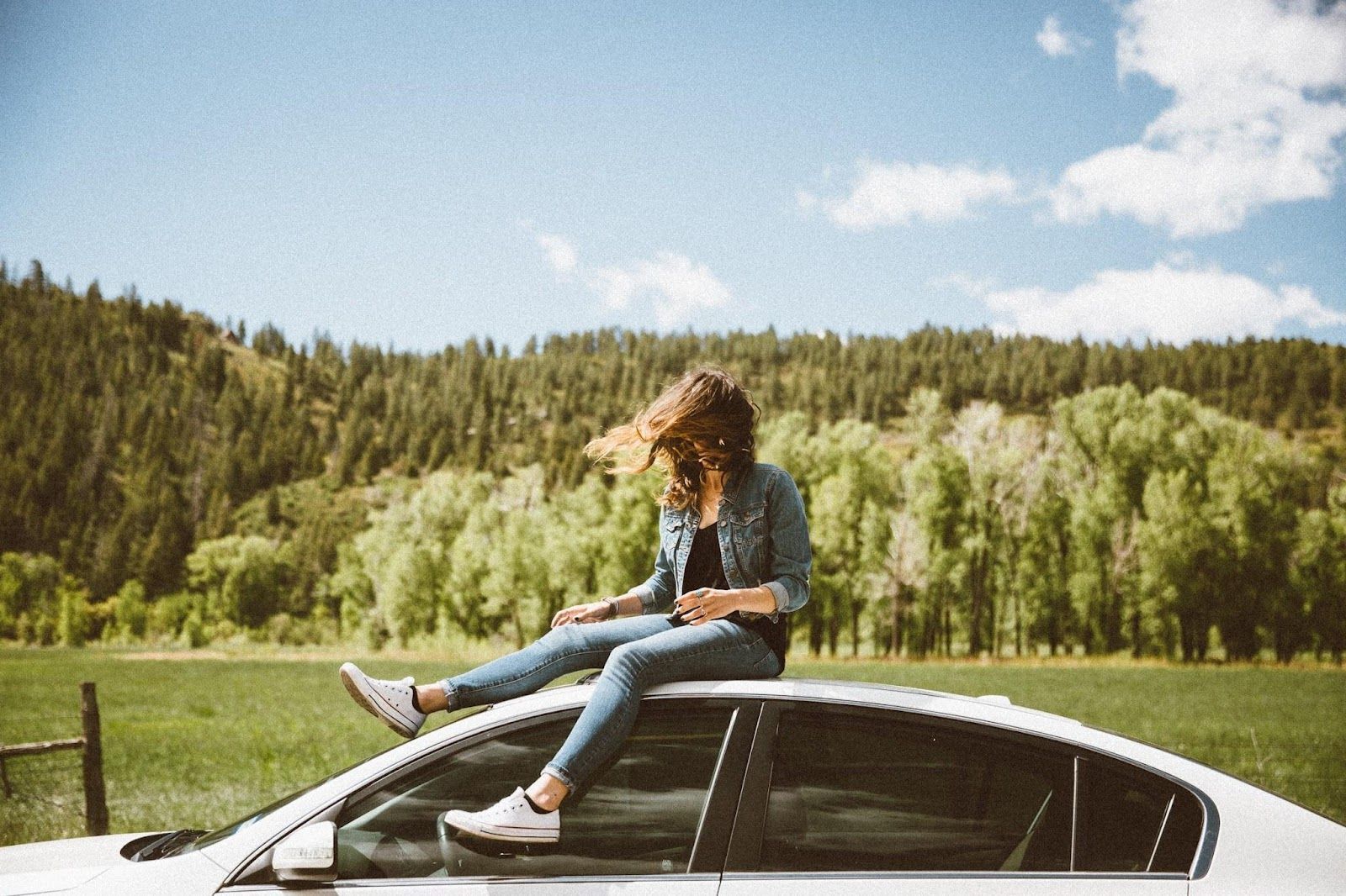 Person sitting on a car roof in a sunny field, with forested hills and blue sky behind.