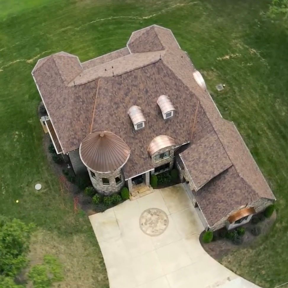 Aerial view of a suburban home with a dark shingled roof, copper accents, a circular tower, and a circular driveway.