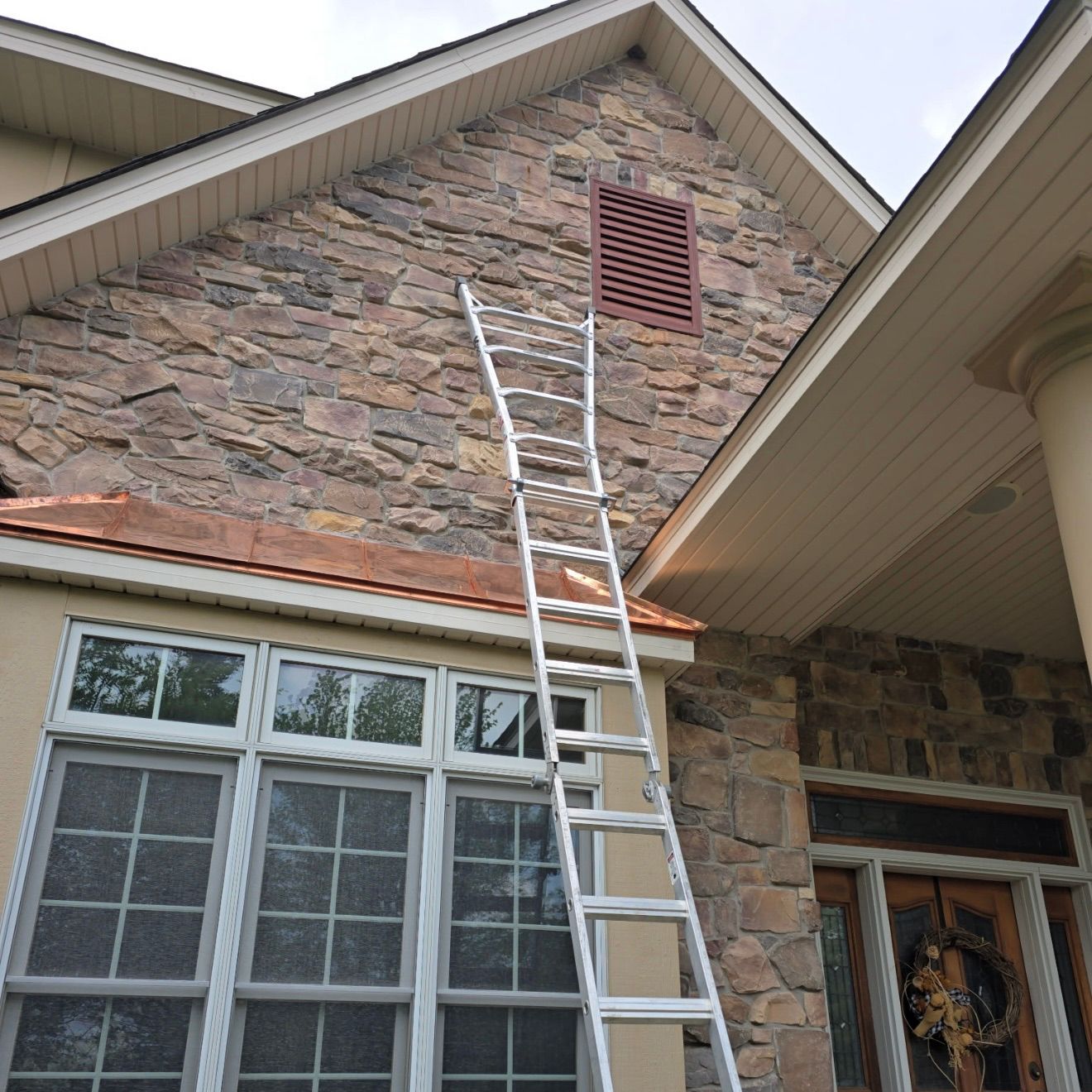 An aluminum extension ladder leans against a stone wall of a house, reaching toward a brown gable vent above a window.