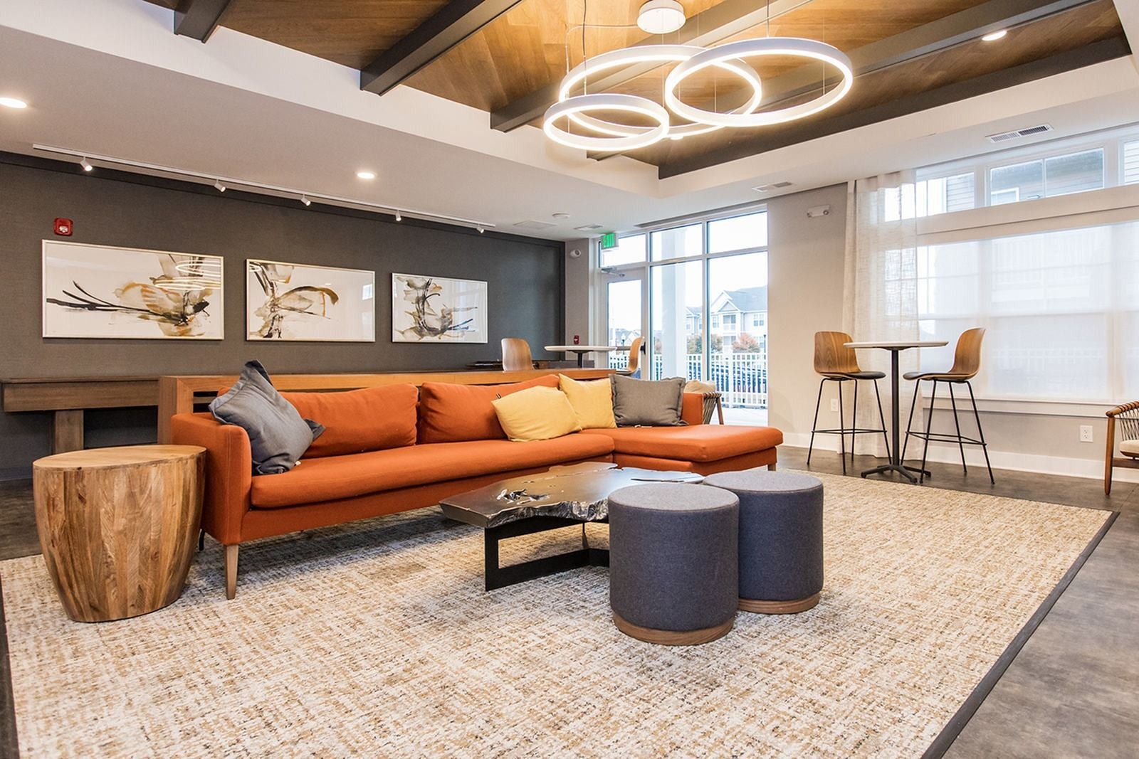 Modern lounge with orange sofa, round ottomans, wood tables, rug, and ceiling lights in a bright office lobby at The Villas at Mill Run Apartments in Owings Mills, MD