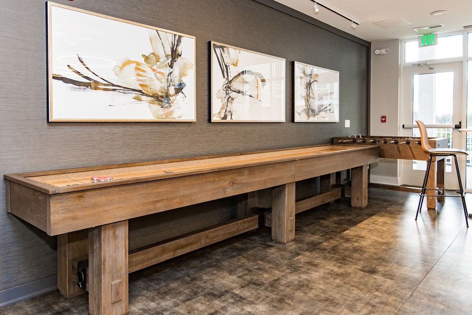 Modern office lounge with a long wooden table, abstract wall art, and stools by a window at The Villas at Mill Run Apartments in Owings Mills, MD