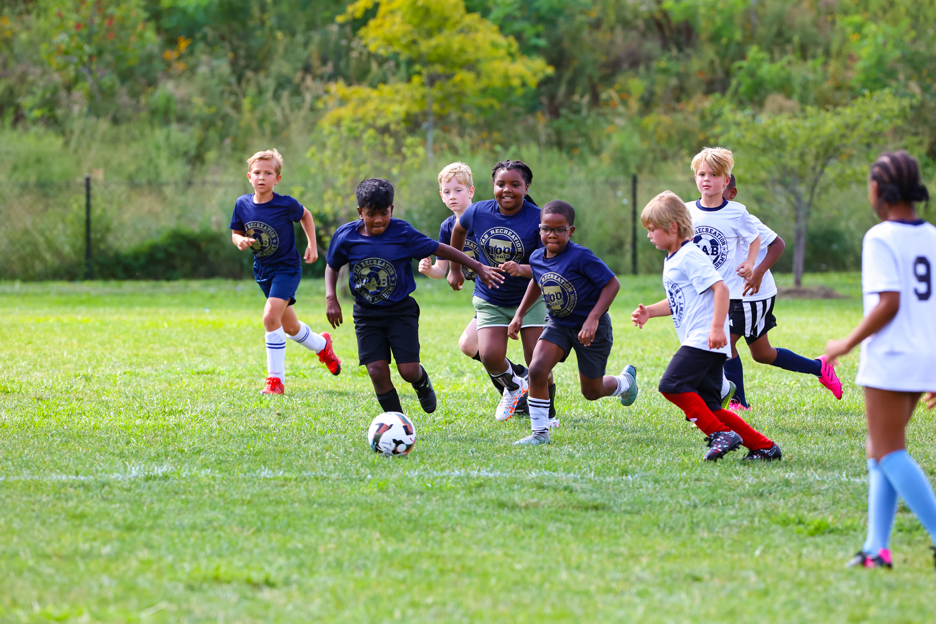 Youth soccer players race after the ball on a grassy field.