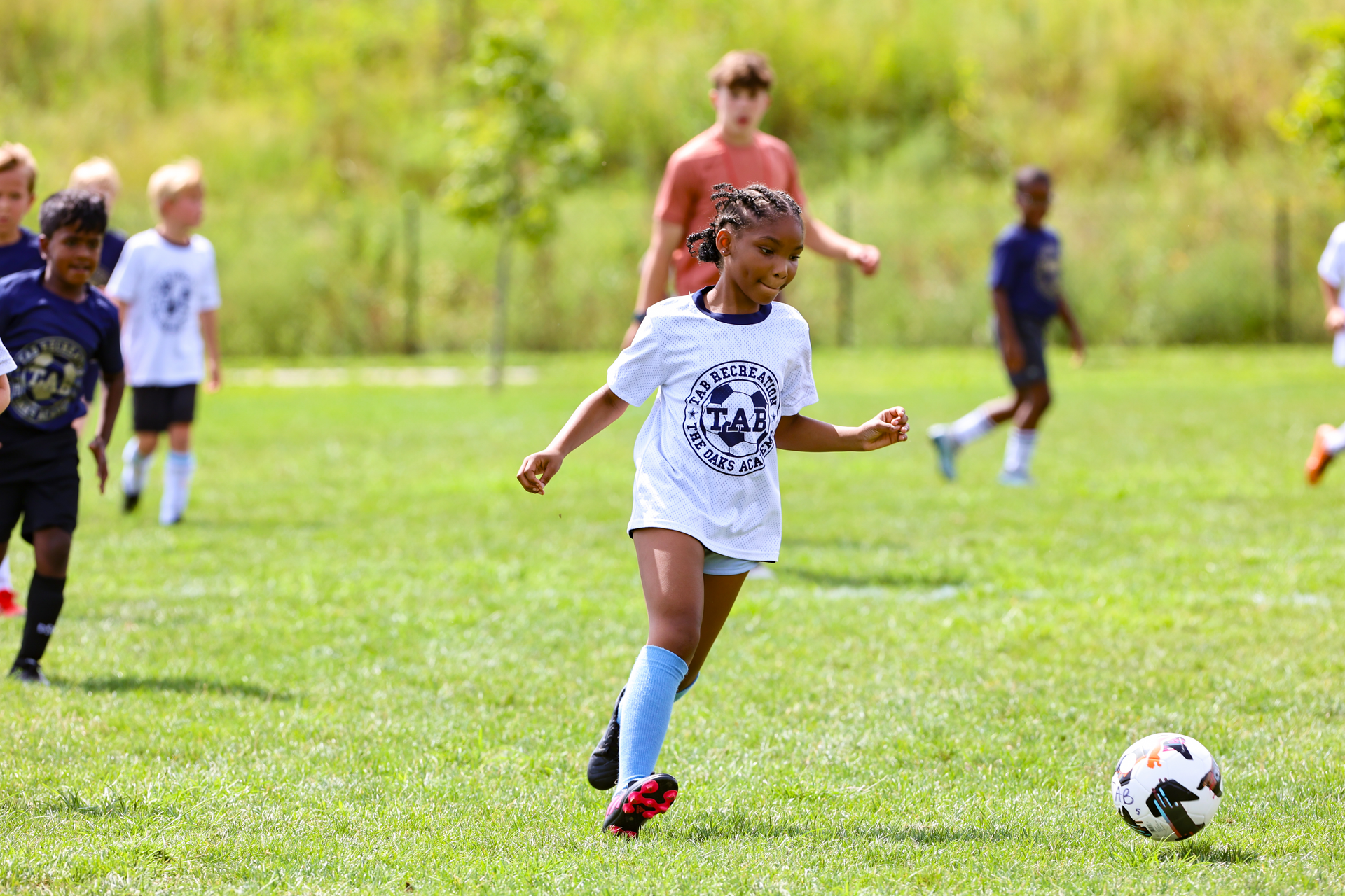 Kids playing soccer on a grassy field, with one player dribbling a ball in the foreground.