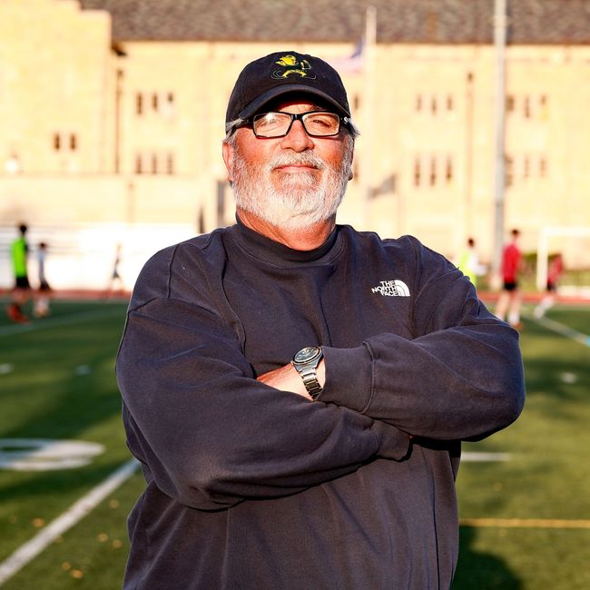 Smiling man in black cap and sweatshirt stands with arms crossed on a football field with a building behind him