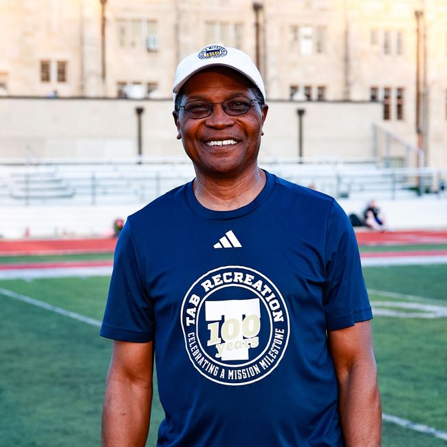 Smiling man in a navy T-shirt and white cap standing on a sports field with stadium steps behind him