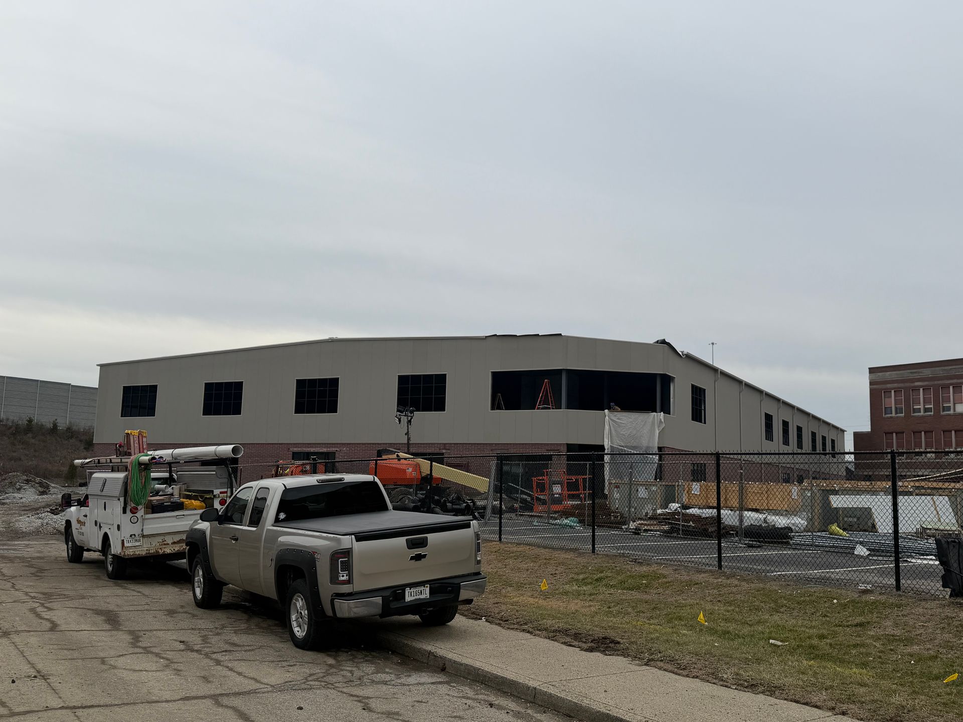 Two utility trucks parked beside a partially built warehouse behind a chain-link fence