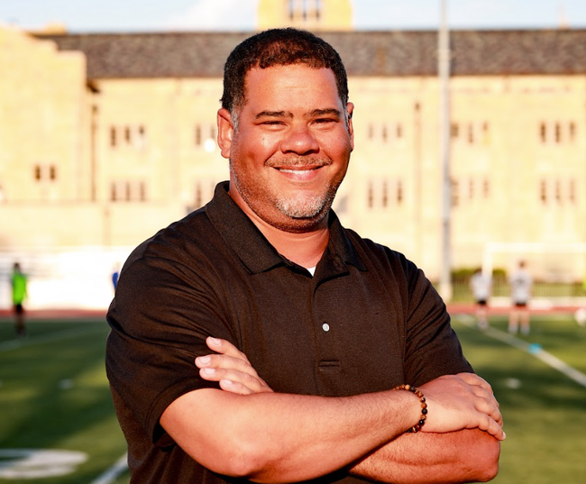 Smiling man with crossed arms on a sports field, wearing a dark shirt at sunset.