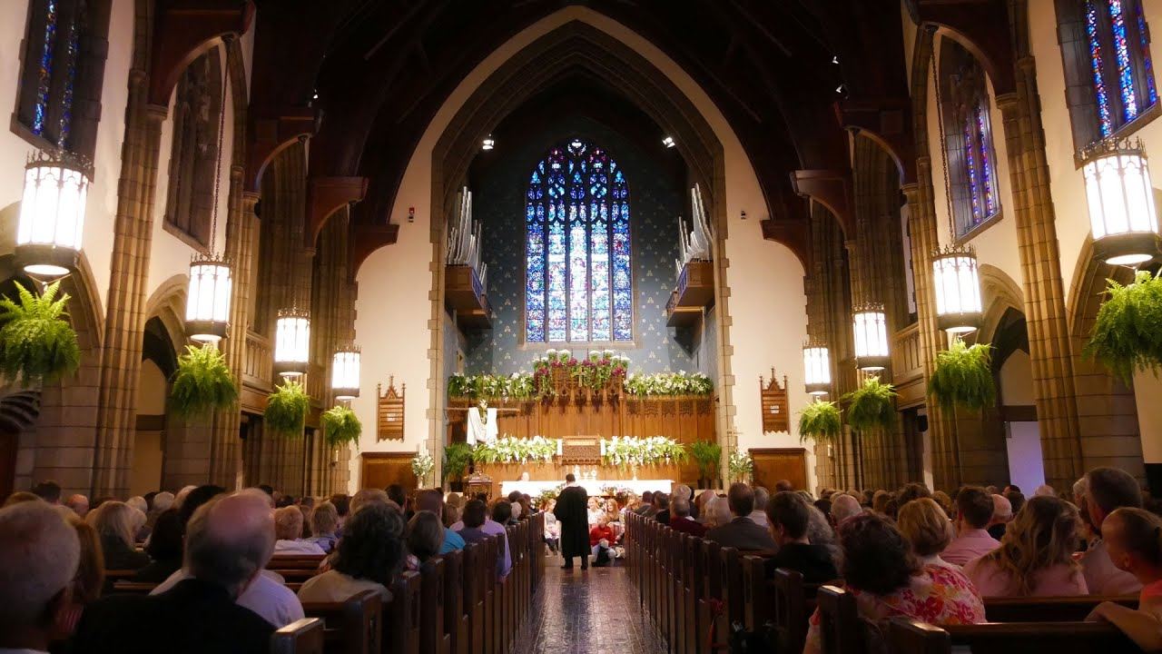 Church wedding ceremony with guests seated in a vaulted nave, altar decorated with flowers and stained glass above.