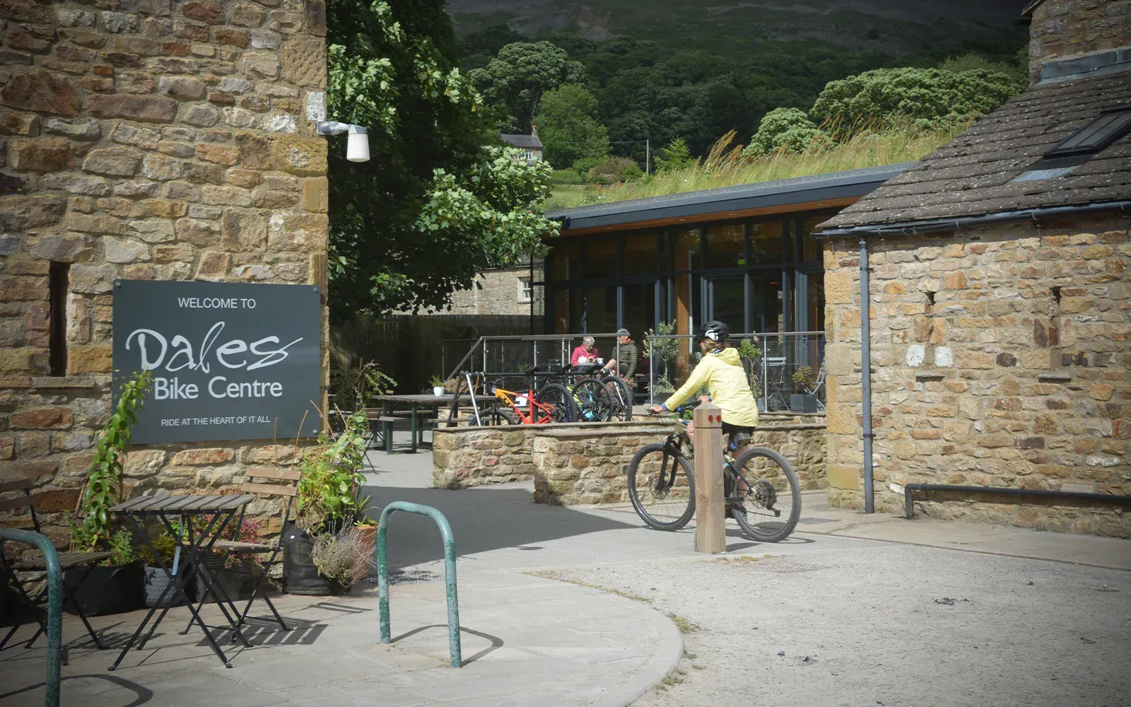 Cyclist passing a stone-walled cafe courtyard with bikes and outdoor tables