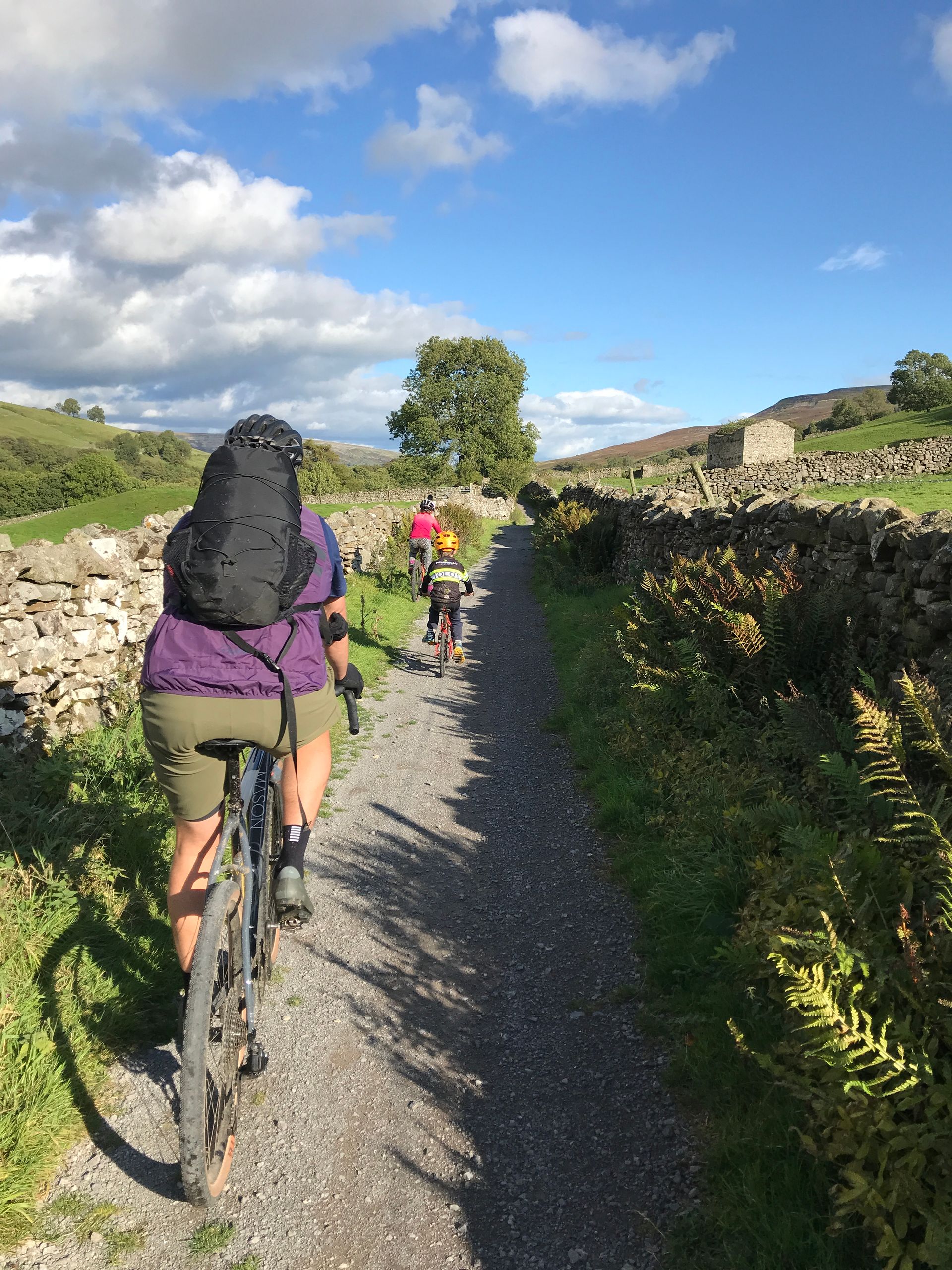 Cyclists riding single file on a gravel trail through green countryside under a blue sky