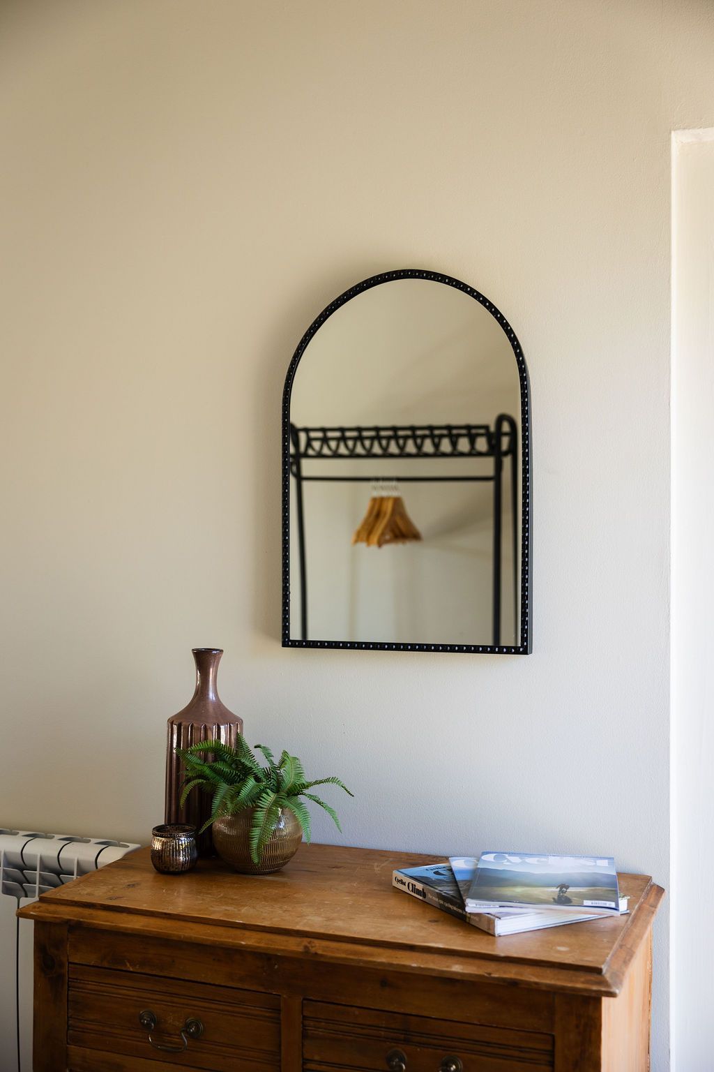 Wall mirror above a wooden dresser with a plant, books, and a decorative vase against a beige wall