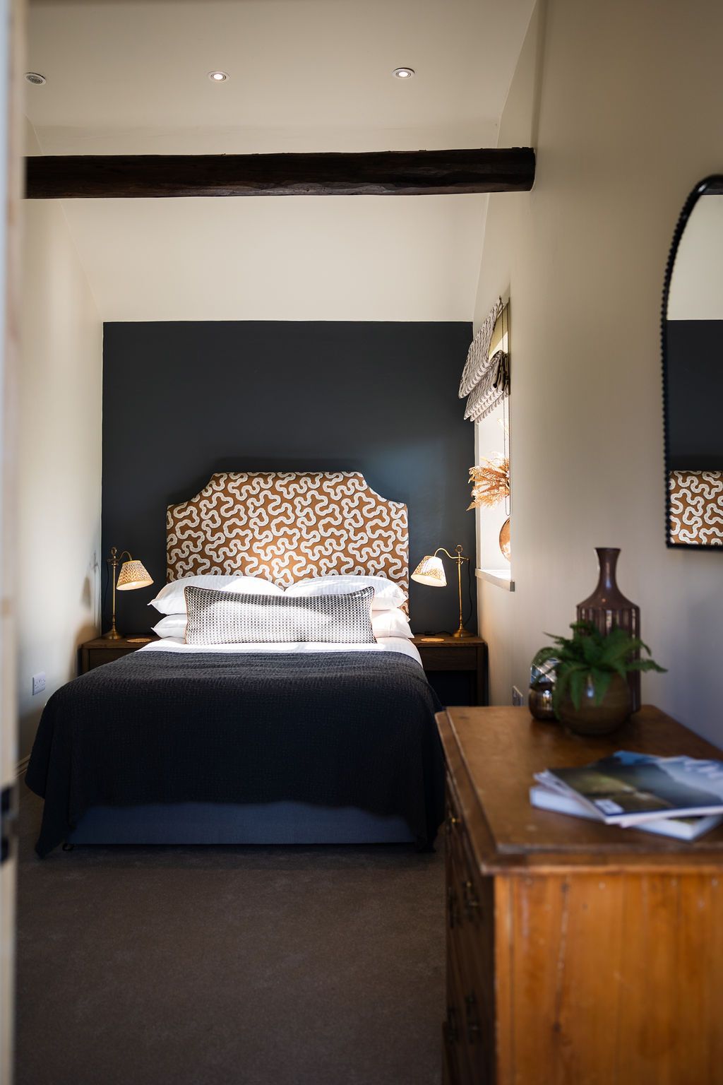 Cozy bedroom with dark accent wall, patterned headboard, neatly made bed, and wooden dresser with flowers