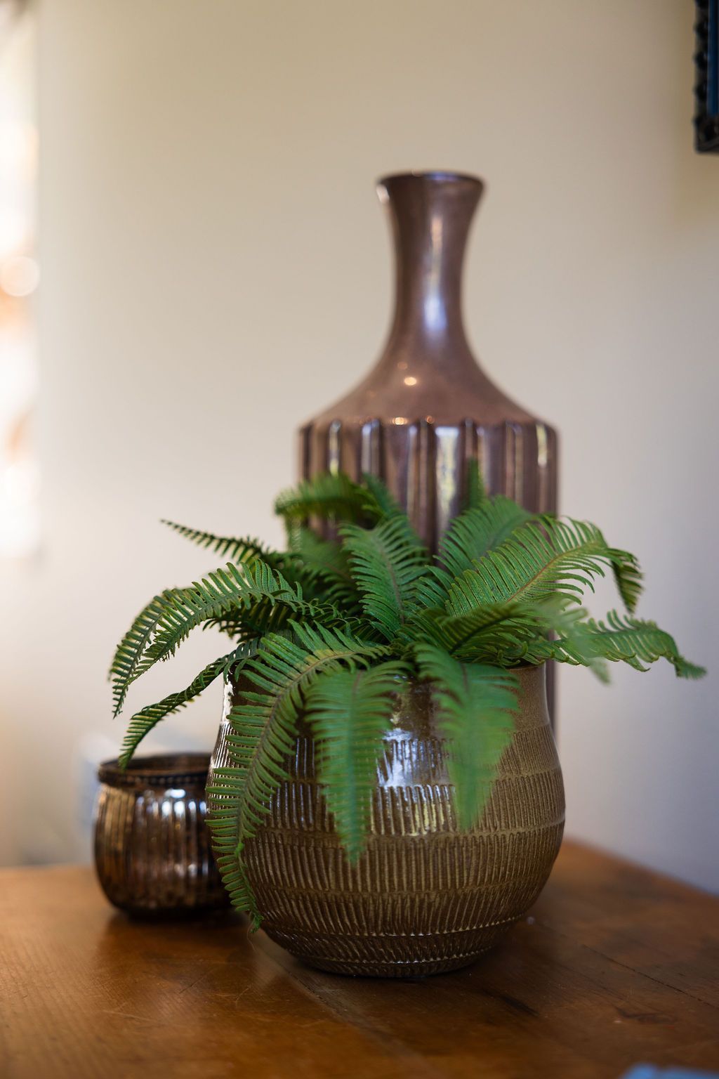 Fern in a woven vase on a wooden table, with a tall brown bottle behind it.