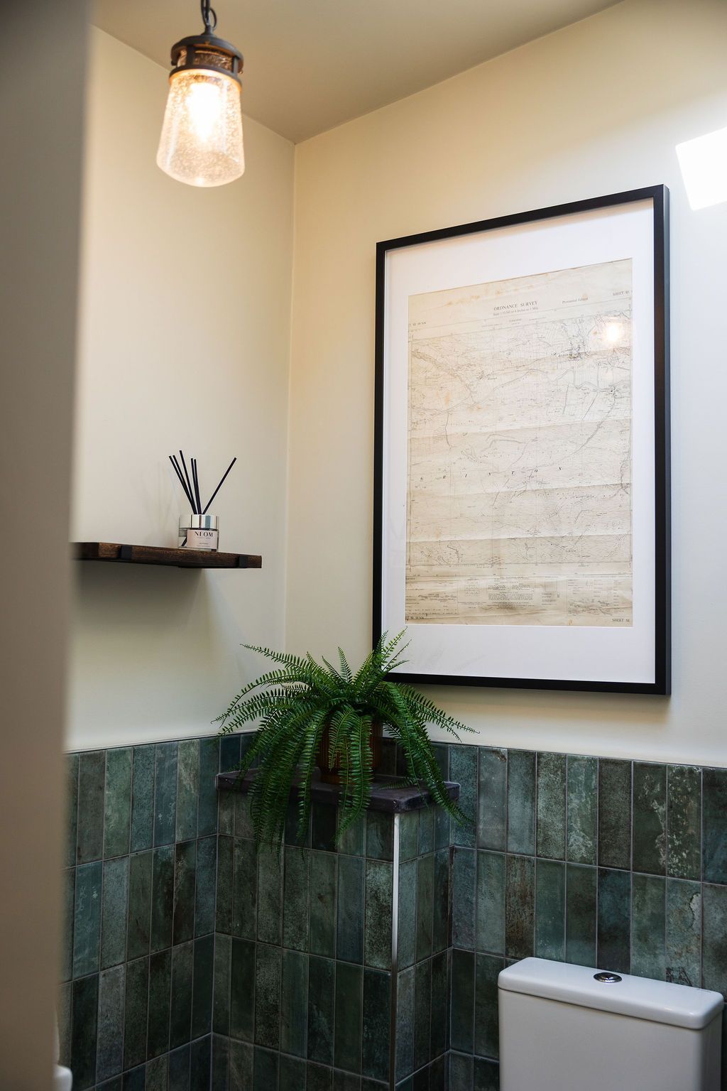 Small bathroom with green tile wainscoting, hanging light, wall shelf, framed art, and a toilet.