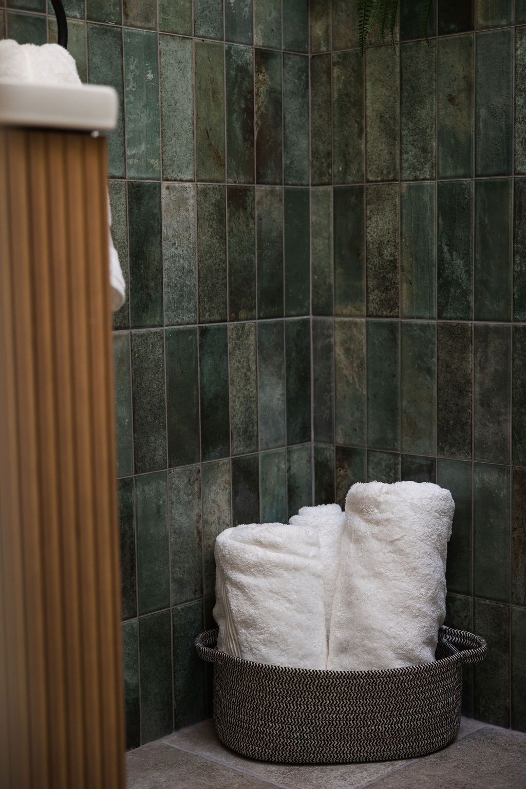 Bathroom corner with green tile wall, wicker basket, and rolled white towels next to a shower curtain.