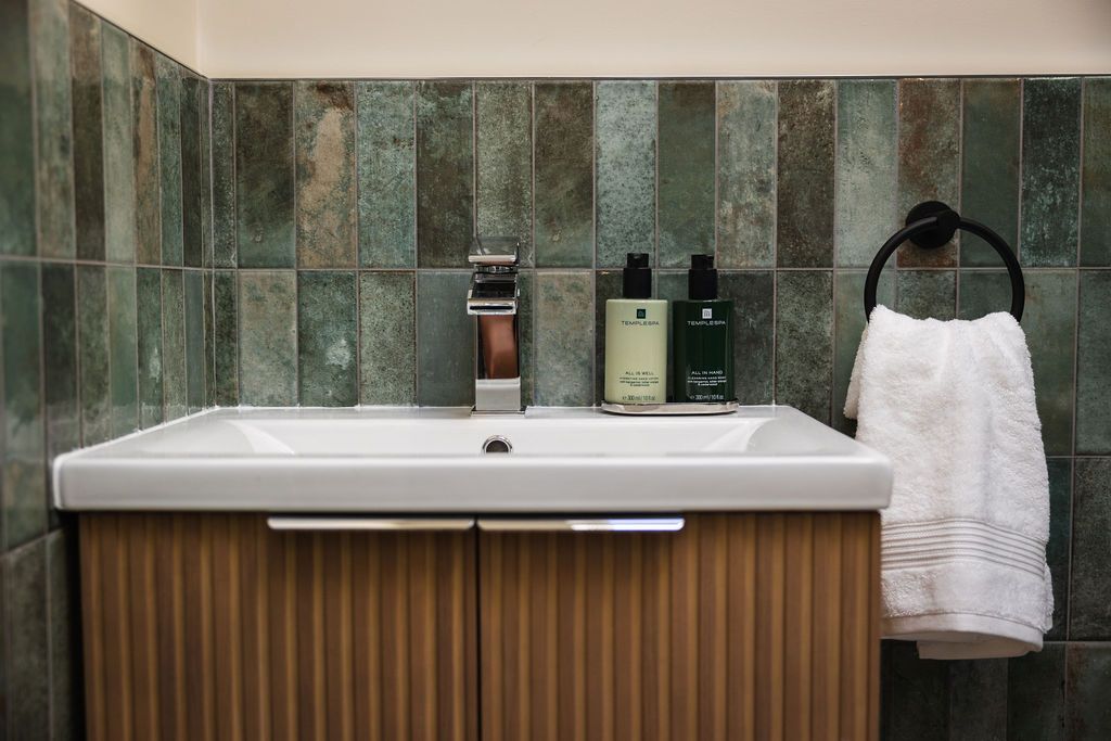 Modern bathroom vanity with white sink, green tile wall, wooden cabinet, soap, and towel rack