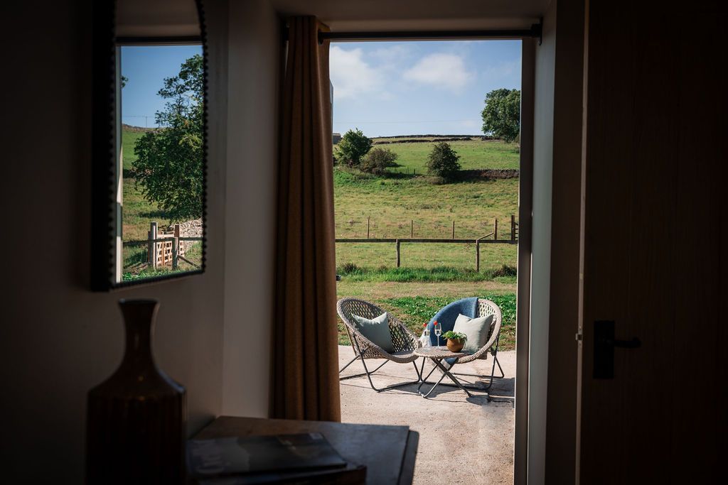 Open doorway to a sunny patio with two chairs, overlooking a fenced green field and trees.