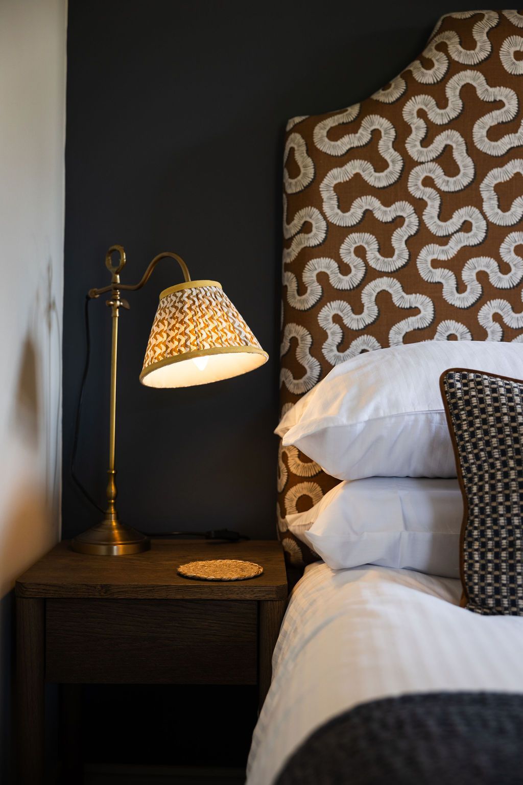Cozy bed with patterned headboard, bedside lamp, and dark wood nightstand in a dim bedroom