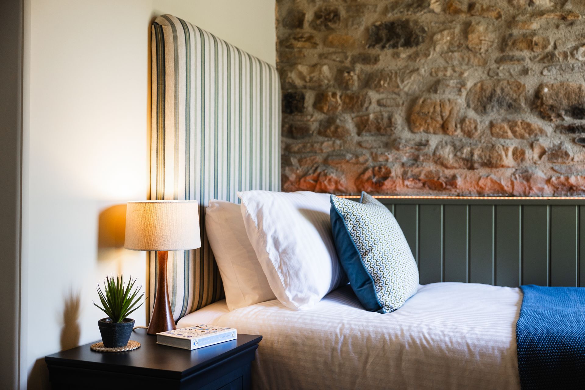Cozy bedroom with striped headboard, white bedding, blue accents, lamp, and rustic stone wall
