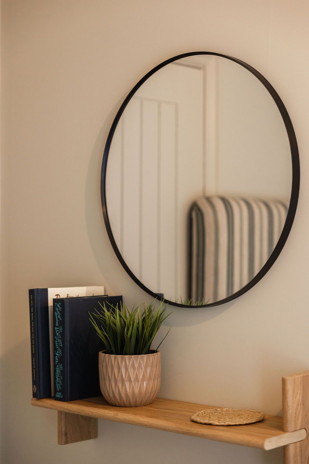 Round wall mirror above a small wooden shelf with books, a potted plant, and a woven coaster