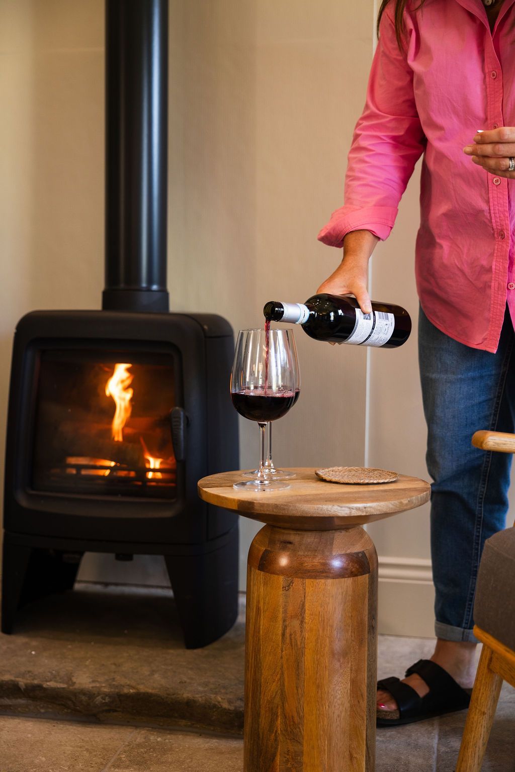 Person pouring red wine into a glass beside a wood stove and small wooden table