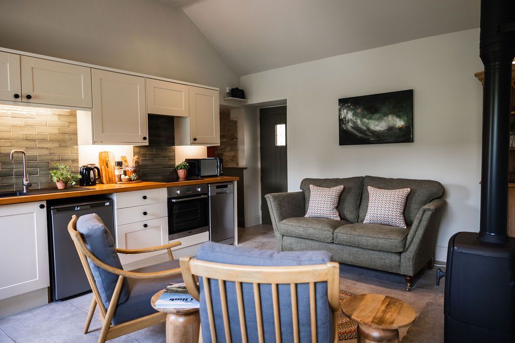 Cozy living room and kitchen with gray sofa, wooden chairs, white cabinets, and a black wood stove