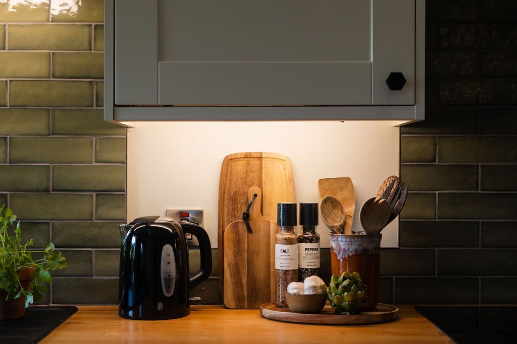 Kitchen counter with kettle, cutting boards, bottles, and potted herbs under warm under-cabinet light